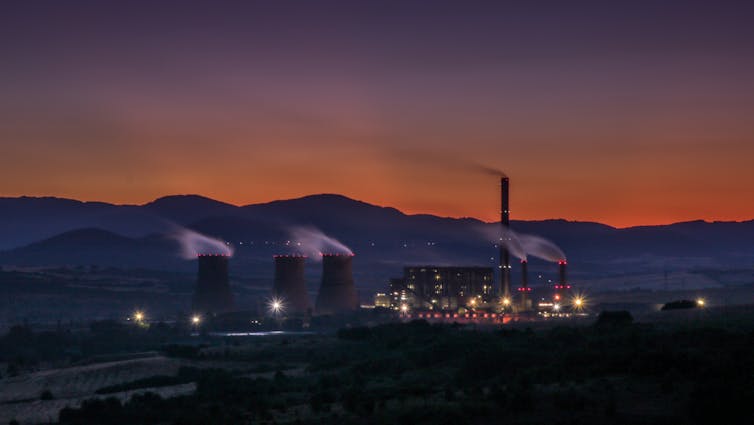 Une usine dans les cheminées fument ; la photo est prise au crépuscule ou à l’aube, avec des montagnes en fond