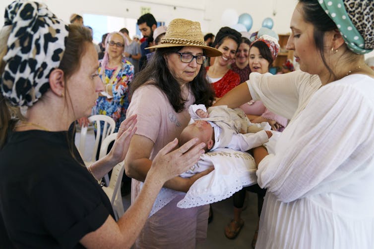 Women arrange a baby on a white pillow one of them is holding, amid a crowd in a large tent.