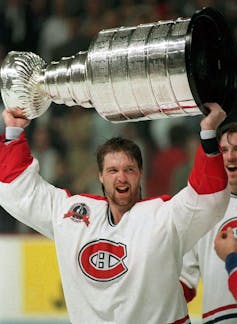 A hockey player in a red and white jersey with the Montréal Canadiens logo in the centre lifts a large, tiered silver trophy over his head.