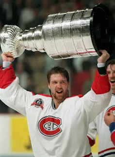 A hockey player in a red and white jersey with the Montréal Canadiens logo in the centre lifts a large, tiered silver trophy over his head.