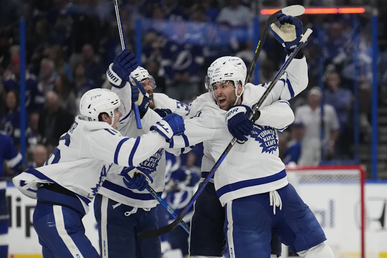 A group of hockey players in blue and white uniforms with the Toronto Maple Leafs logo on them hug each other in celebration