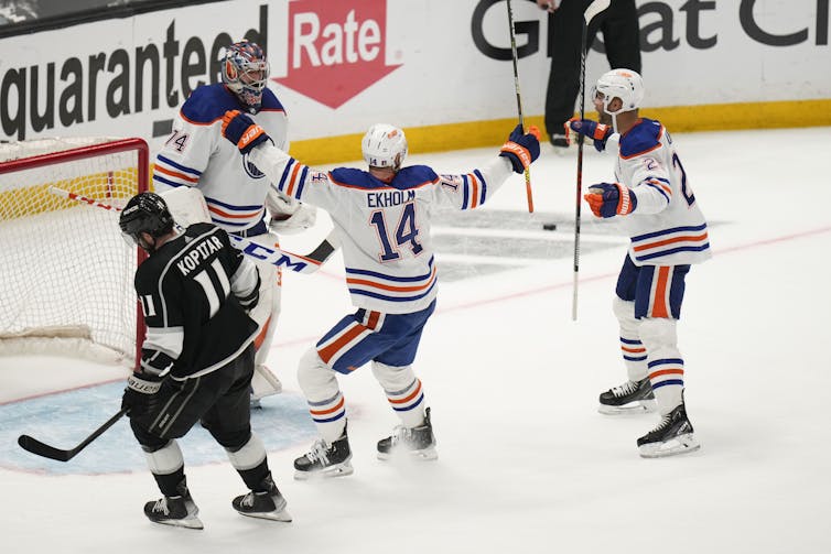 A group of hockey players in white, blue and orange uniforms skate towards each other with their arms out. A hockey player in black and white skates by them with his head down.