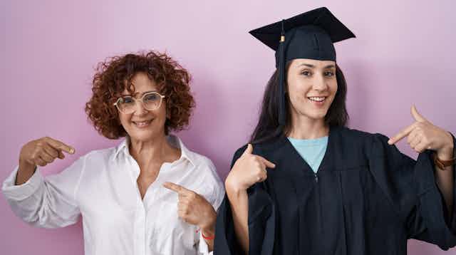 A graduate seen in a cap pointing to herself next to a middle-aged woman in a white shirt pointing to herself.