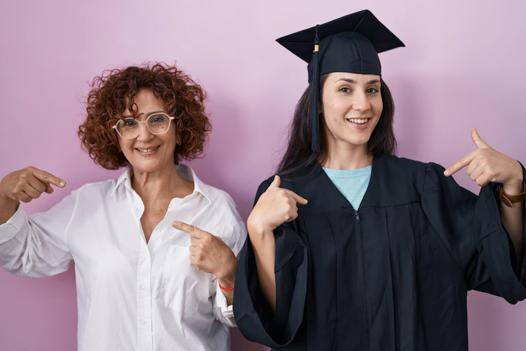 A graduate seen in a cap pointing to herself next to a middle-aged woman in a white shirt pointing to herself.