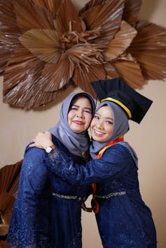Two women seen embracing, one in a graduation cap, both wearing headscarves.