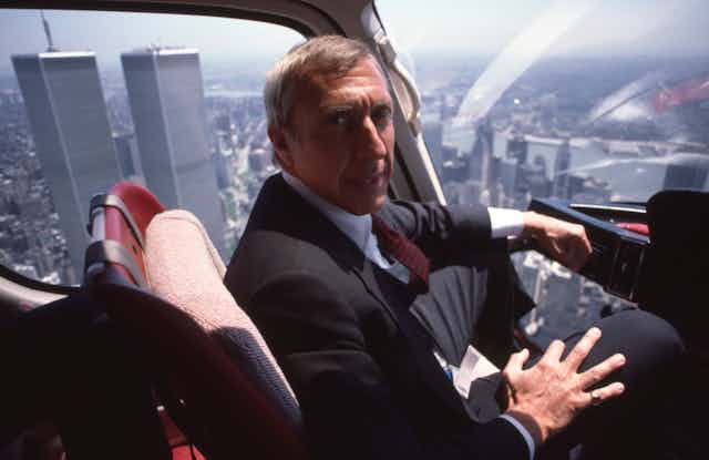 an older man in a suit rides in an aircraft over Manhattan
