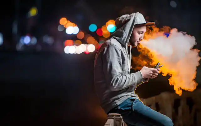 Young man vapes outside, looking at his phone