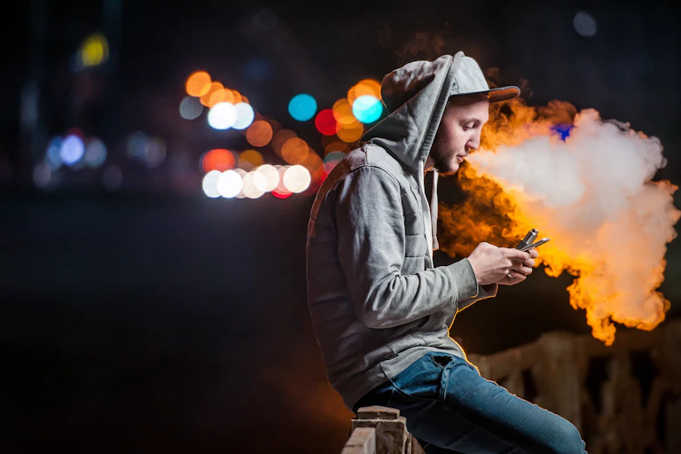 Young man vapes outside, looking at his phone