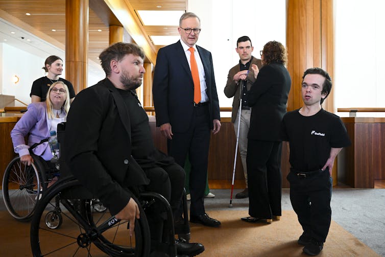 man in wheelchair in centre talks to man of small stature, man in red tie stands behind him
