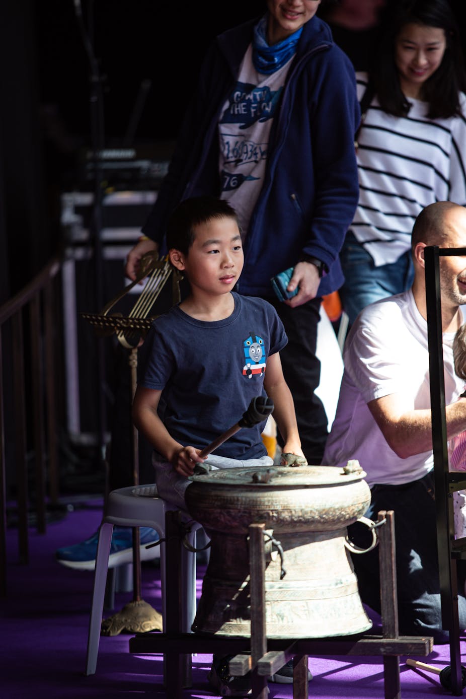 Child holding a rubber striker and sitting next to a bronze drum in front of other audience members 