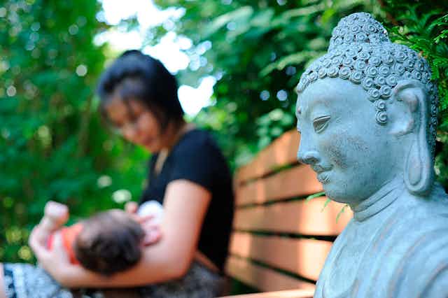 A blurry photo shows a woman in a black shirt nursing a child on a bench, with a statue of the Buddha nearby.