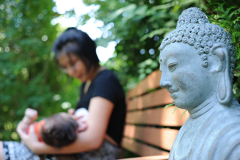 A blurry photo shows a woman in a black shirt nursing a child on a bench, with a statue of the Buddha nearby.