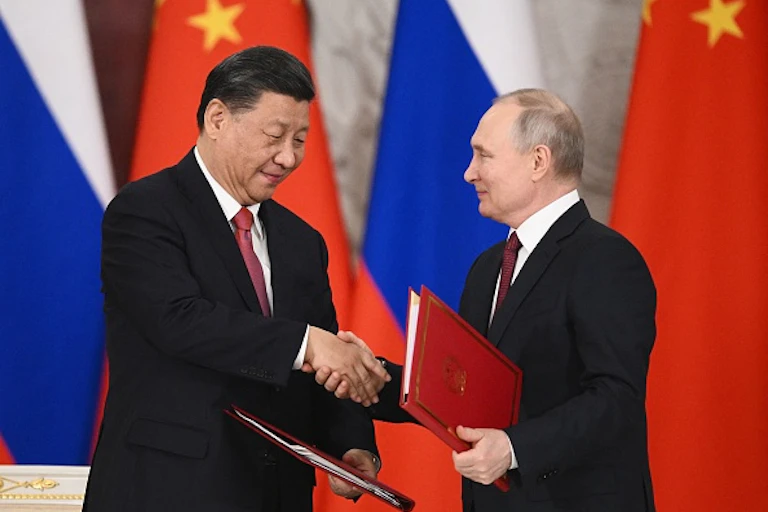 Two men wearing suits shake hands and exchange documents.