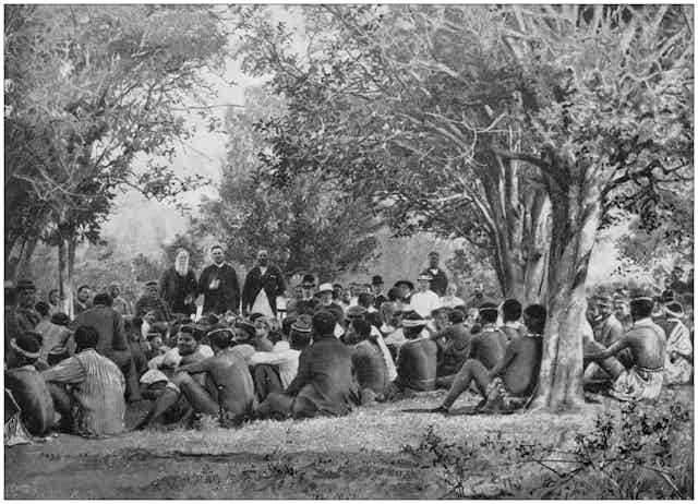 A group of Zulu people sit on the ground underneath trees looking at three men who stand addressing them, dressed in church clothes, one holding a book under his arm.