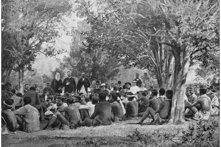 A group of Zulu people sit on the ground underneath trees looking at three men who stand addressing them, dressed in church clothes, one holding a book under his arm.