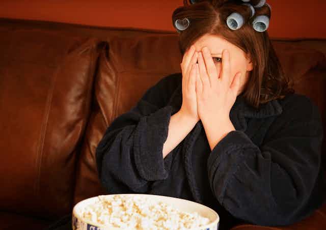 Woman with curlers in her hair watching TV covering her face with her hands.