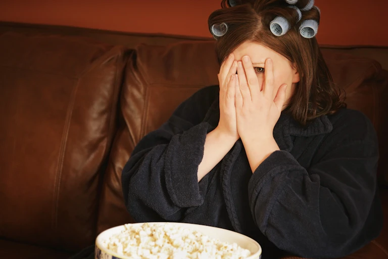 Woman with curlers in her hair watching TV covering her face with her hands.