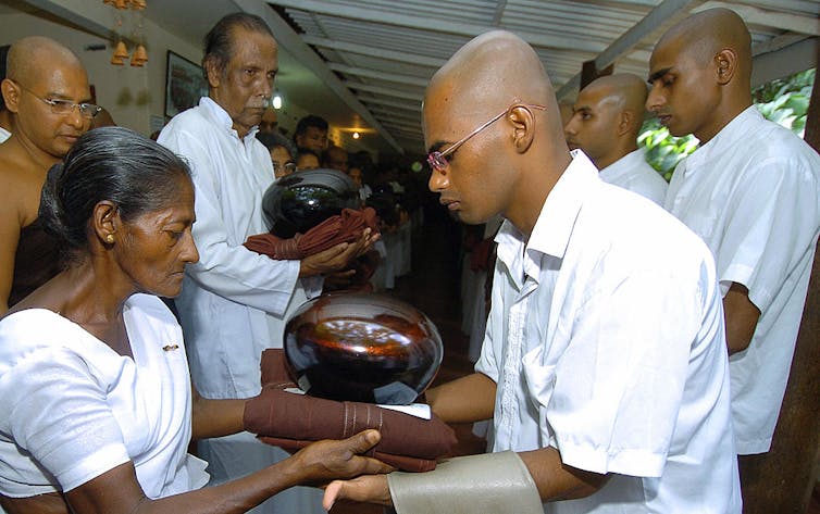 A woman in a simple white shirt ceremonially hands a rolled piece of fabric to a young man with a shaved head in a white shirt.