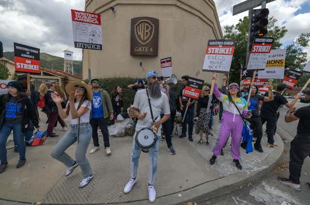 Trabajadores en huelga con pancartas, cantando y bailando frente a un estudio cinematográfico.