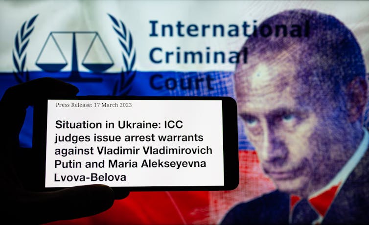 The silhouette of a phone shows the words, 'Situation in Ukraine: ICC judges issue arrest warrants against Vladimir Vladimirovich Putin and Maria Alekseyevna Lvova/Belova' against a white, blue and red stripped backdrop, with a man's face on the right side of it.