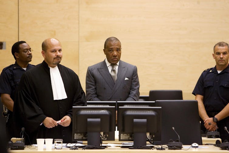 A Black man in a grey suit looks at the camera, surrounded by someone in a judge's black robe and what appear to be security guards in navy outfits.