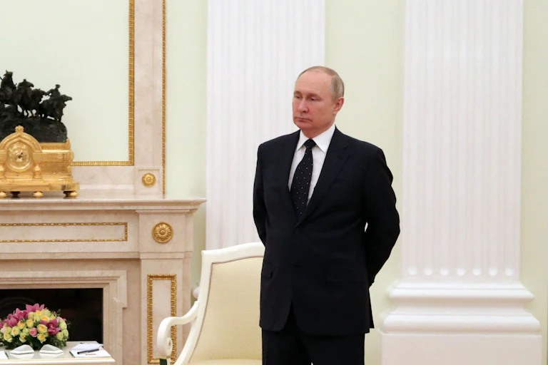 A man in a black suit stands in a formal looking room with two chairs.