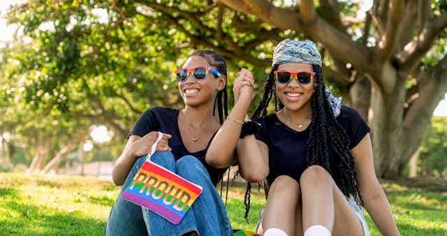 Two young Black women wearing rainbow-colored sunglasses hold hands and smile in park