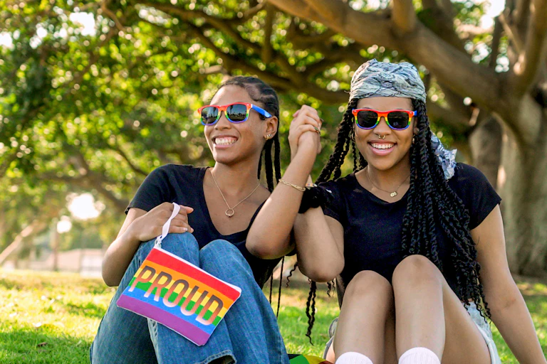Two young Black women wearing rainbow-colored sunglasses hold hands and smile in park
