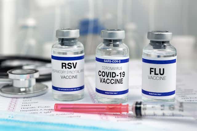 Vials of RSV, COVID-19 and flu vaccines are on a table. Two syringes are in the foreground.