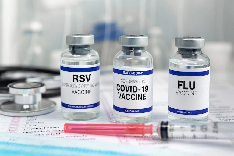 Vials of RSV, COVID-19 and flu vaccines are on a table. Two syringes are in the foreground.