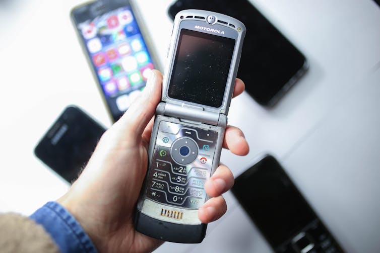 A handing holding a flip cellphone over a table covered with an assortment of smartphones.