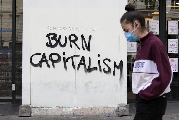 A sign reads Burn Capitalism during a protest. A woman wearing a mask stands next to the sign.