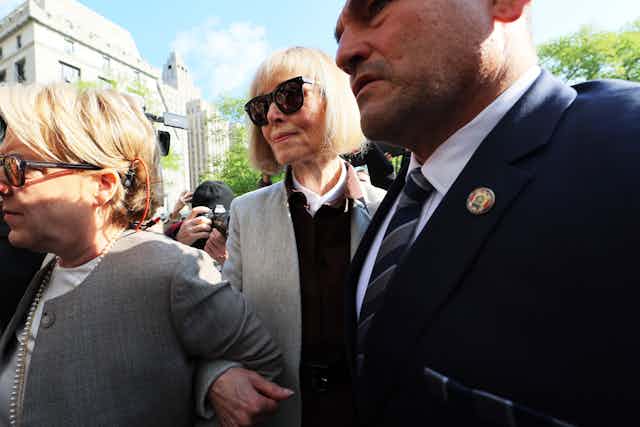 A woman with short white hair an black glasses wears a blazer and walks between two people outside.