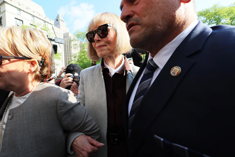 A woman with short white hair an black glasses wears a blazer and walks between two people outside.