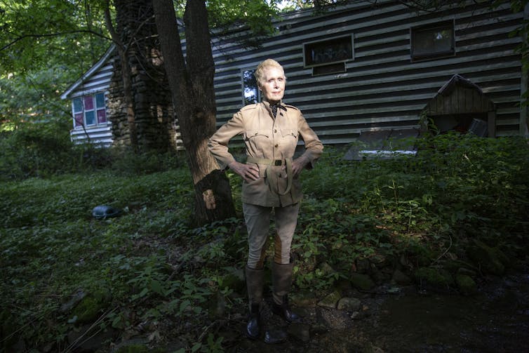 An older woman wears a beige outfit and stands in the woods in front of a low lying house.