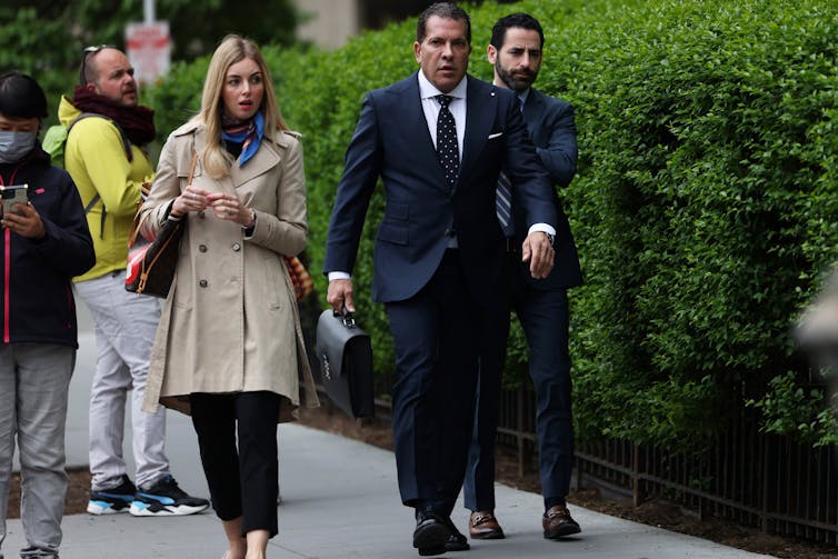 Un hombre blanco con un traje azul oscuro camina por una acera, flanqueado por una mujer con una chaqueta beige y otro hombre con un traje.