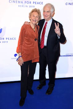An older man in a suit flashes a peace sign, one arm around an older woman in an orange top and black slacks.