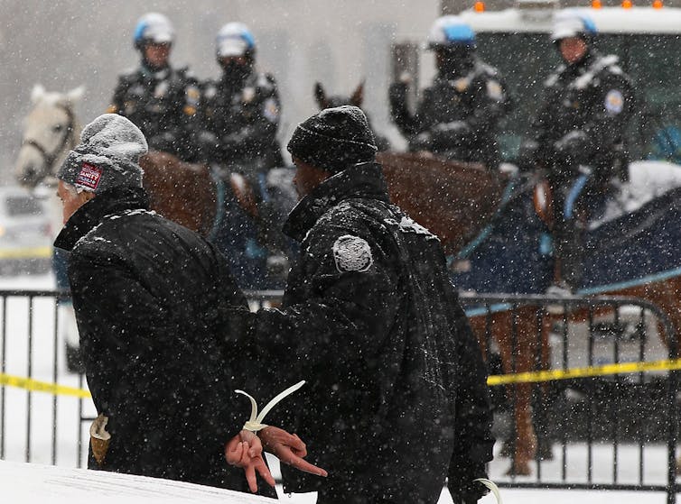On a snowy day, a police officer escorts a man whose hands are in zip ties, as police on horses look on.