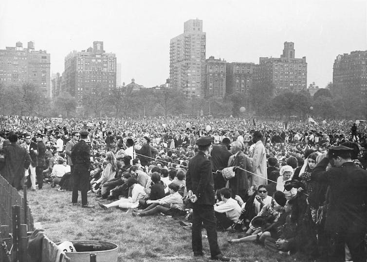 A black and white photo of a huge crowd seated in a park, with skyscrapers in the background.