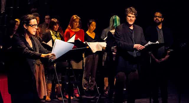 Actors seen standing on a stage holding scripts against a black background.