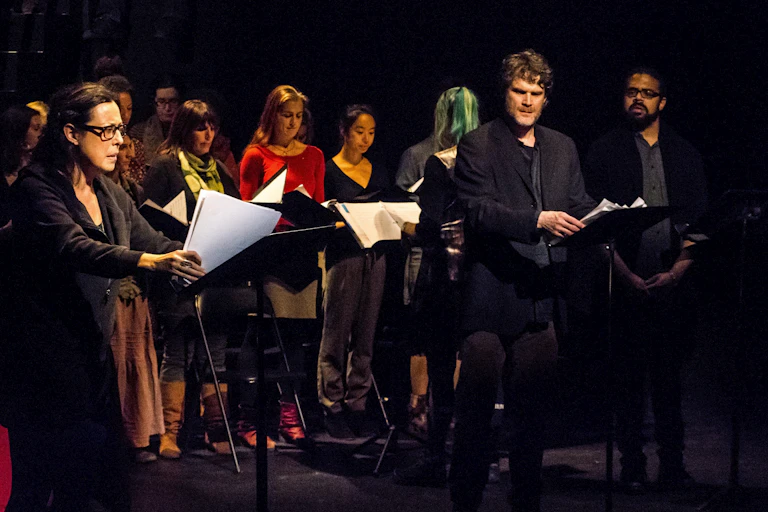 Actors seen standing on a stage holding scripts against a black background.