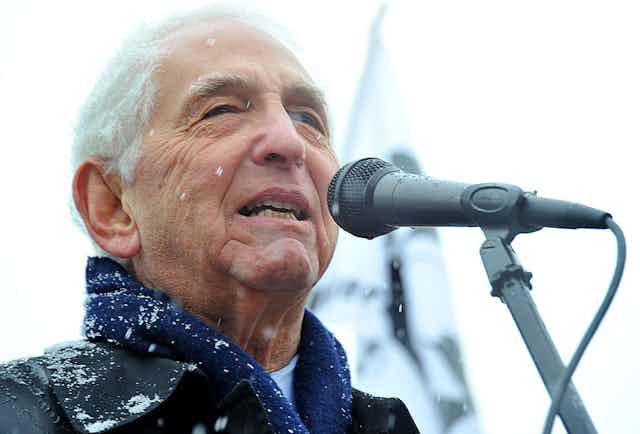 A white-haired man in a winter jacket speaks into a microphone in a close-up photo.