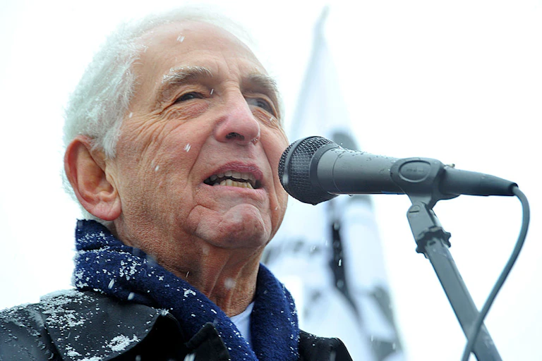 A white-haired man in a winter jacket speaks into a microphone in a close-up photo.