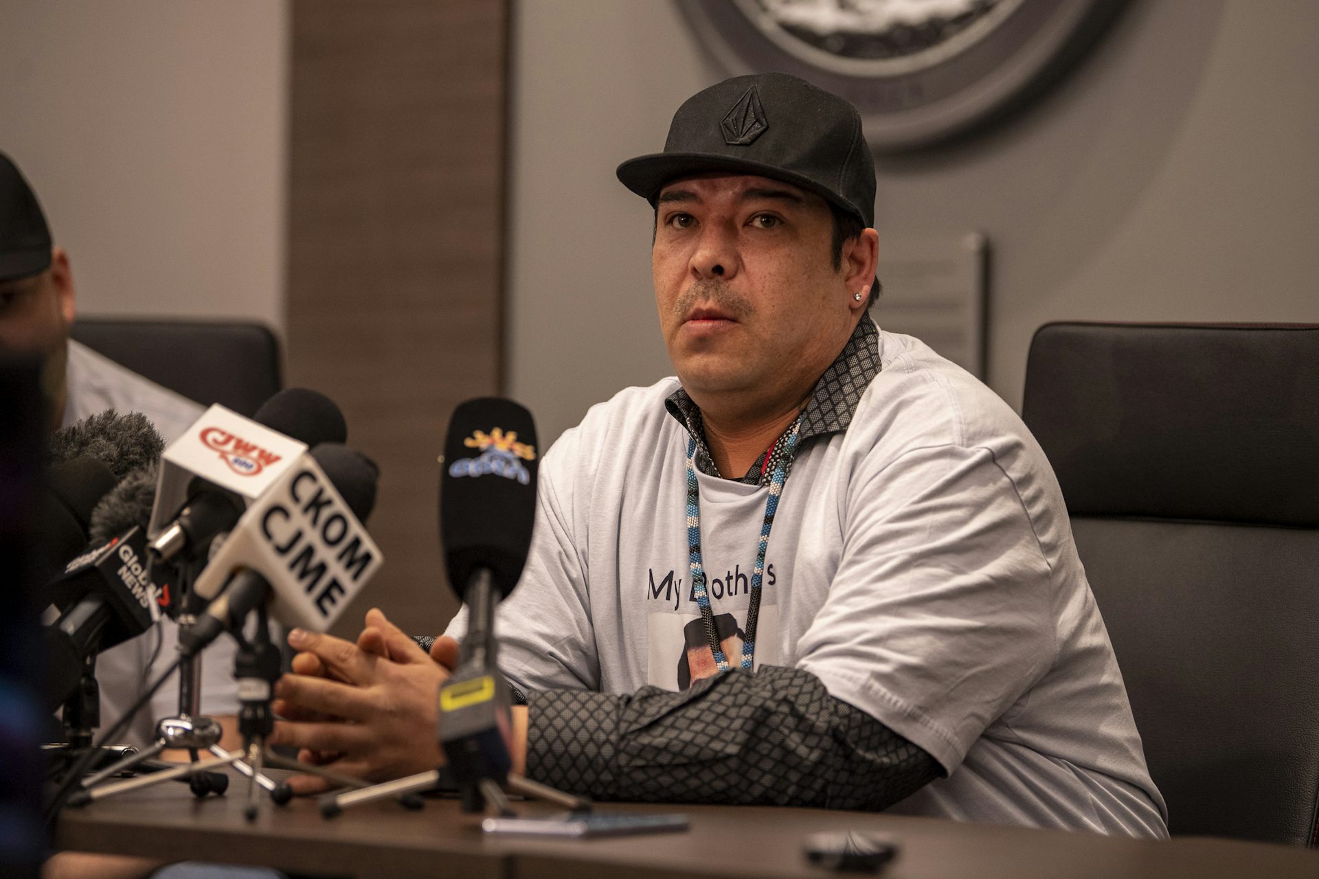 A man in a white T-shirt and black ball cap speaks into a microphone.