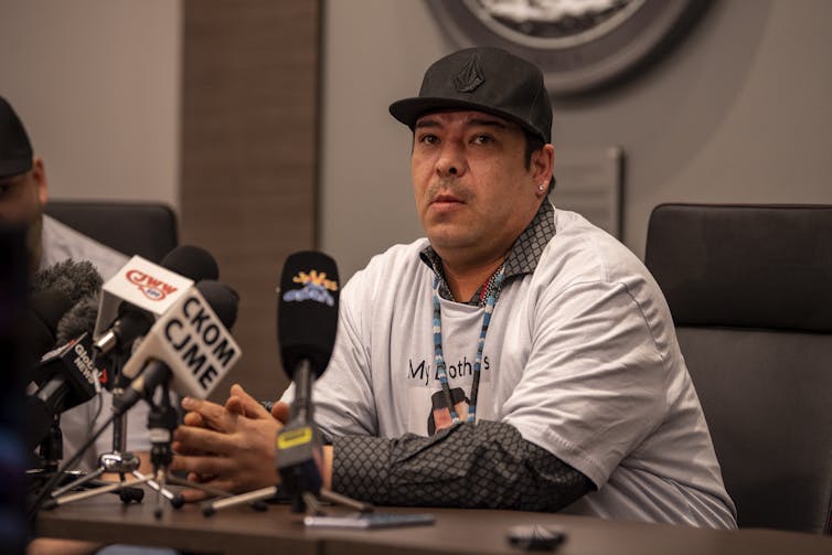 A man in a white T-shirt and black ball cap speaks into a microphone.