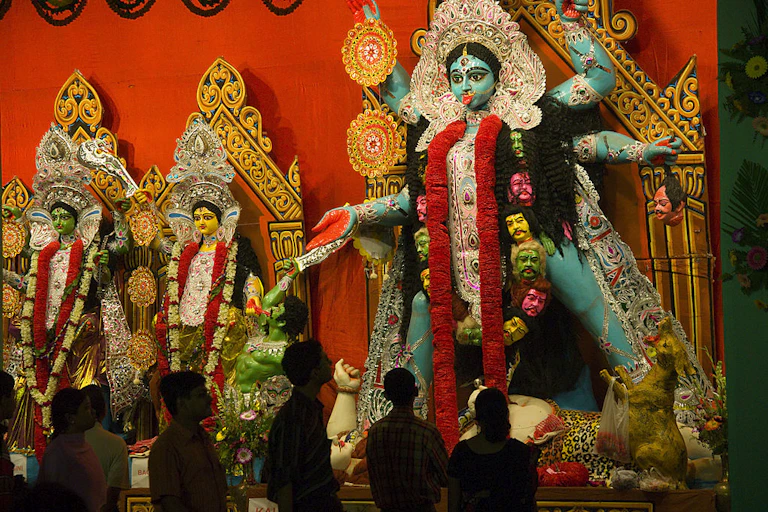 A brightly colored teal statue of a woman with many arms and legs displayed against a bright-red wall.