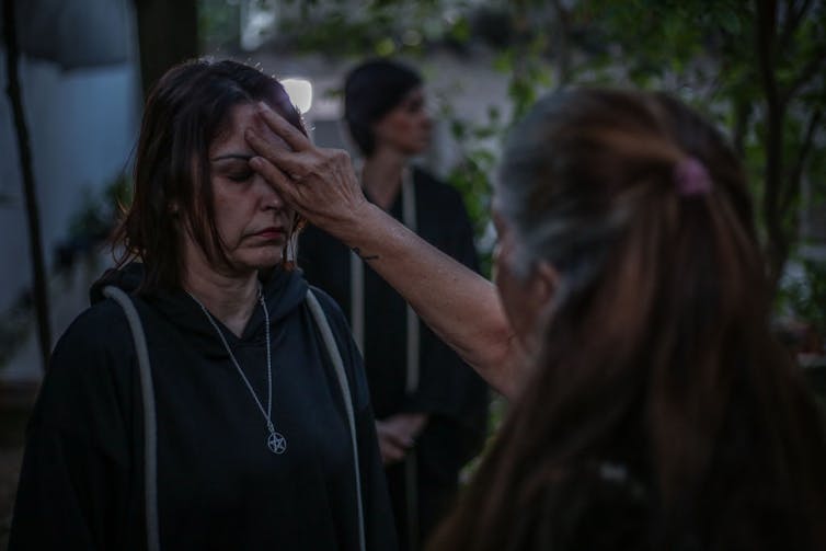 A woman wearing dark clothing smudges a substance on the forehead of another woman with her eyes closed.