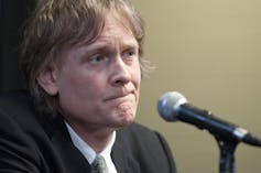 A shaggy-haired blonde man with a microphone in front of him.