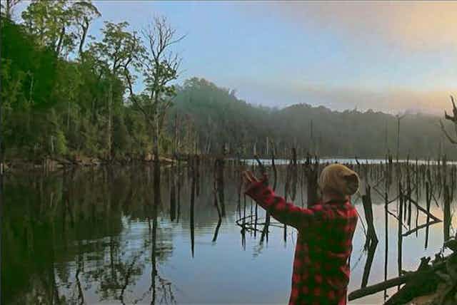 Man in red check flannel shirt and beanie raising arms as he stands next to a lake