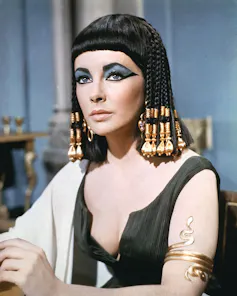 A woman looks wryly ahead, dressed in a low-cut outfit with a beaded headpiece and prominent eyeshadow on.
